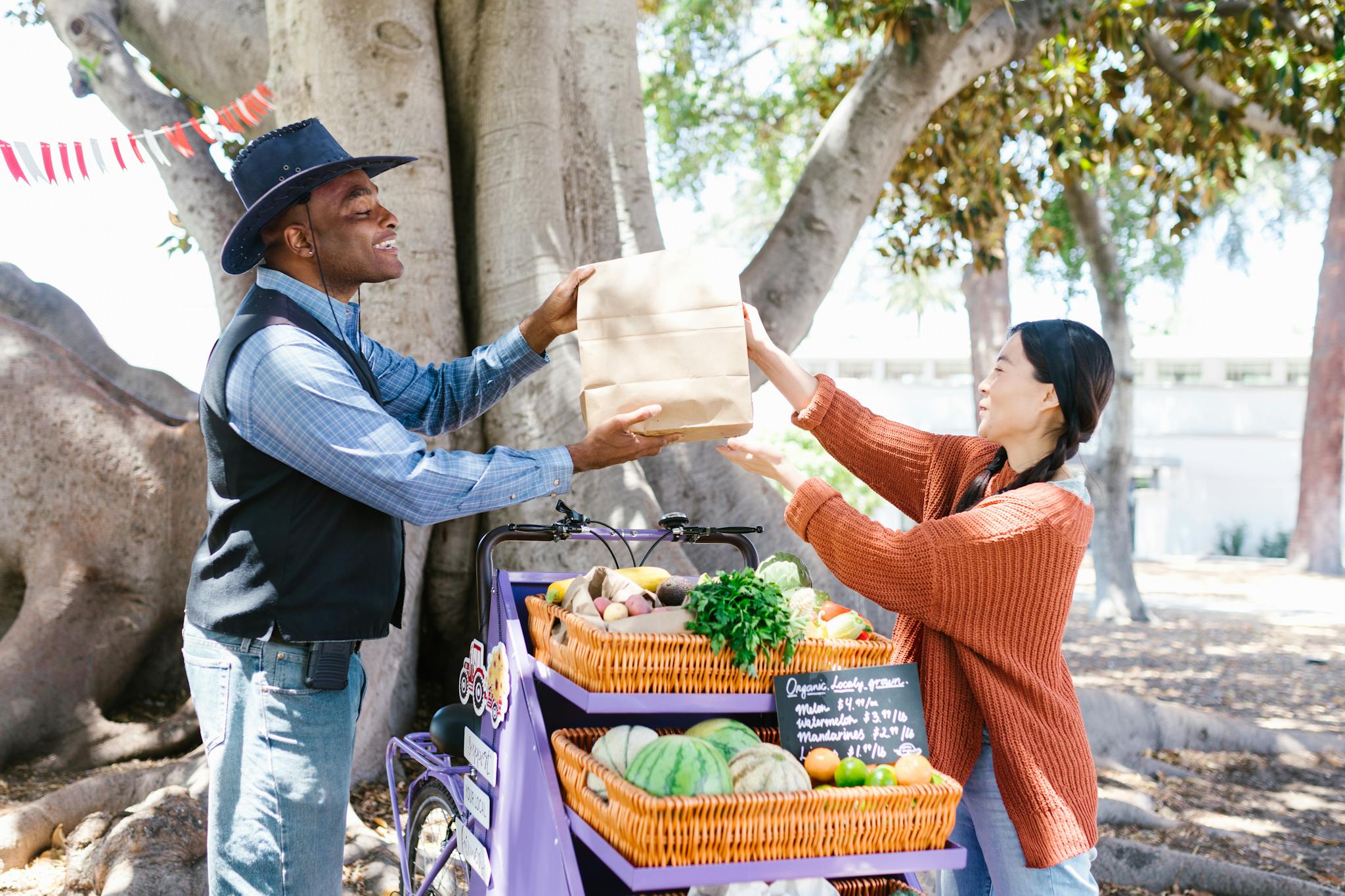 A vendor hands a paper bag to a customer at an outdoor farmer's market filled with fresh produce.